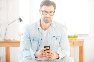 My work is my life. Handsome young man in shirt and eyewear holding mobile phone and looking at camera while sitting in front of his working place photo