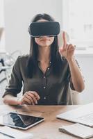 It seems you can touch it. Confident young woman in virtual reality headset pointing in the air while sitting at her working place in office photo