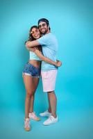 Full length of beautiful young couple embracing and smiling while standing against blue background photo