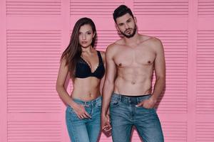 Made to love each other. Beautiful young couple holding hands and looking at camera while standing against pink background photo