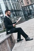 enterado de las últimas noticias. un joven apuesto con traje completo leyendo un periódico mientras se sienta al aire libre foto