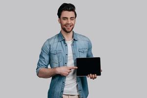 Copy space on his tablet. Handsome man in blue jeans shirt pointing copy space on his digital tablet and looking at camera while standing against grey background photo