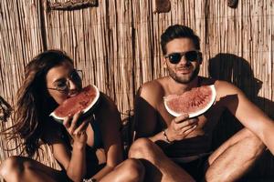 Enjoying summer refreshments. Beautiful young couple eating watermelon and smiling while sitting outdoors photo