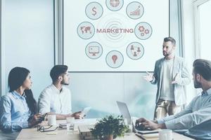 Discussing marketing strategy. Group of business people sitting at the office desk together while their colleague telling something and gesturing with large white board in the background photo