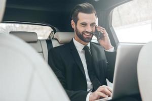 centrándose en el trabajo. apuesto joven hombre de negocios trabajando en su computadora portátil y hablando por teléfono mientras está sentado en el asiento trasero del camión foto