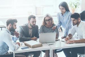 Discussing business issues. Group of young business people sitting at the office desk and discussing something while looking at laptop together photo