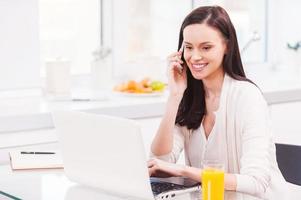 Having opportunity to work everywhere. Attractive young woman working on laptop and talking on the mobile phone while sitting in the kitchen photo