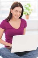 Surfing the net at home. Attractive young woman working on laptop and smiling while sitting on the floor at her apartment photo