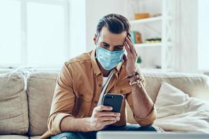Thoughtful young man in protective face mask using smart phone while spending time at home photo