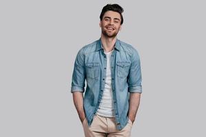 Perfect man. Handsome young man in blue jeans shirt keeping hands in pockets and looking at camera while standing against grey background photo