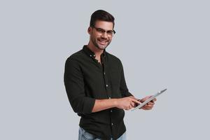 Always ready to help. Good looking young man in eyeglasses holding his digital tablet and looking at camera with smile while standing against grey background photo