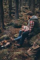 Traveling with beloved. Beautiful young couple holding cups while warming up near the campfire photo