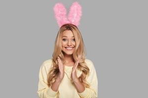 Excited young smiling woman in pink bunny ears clapping and looking at camera while standing against grey background photo