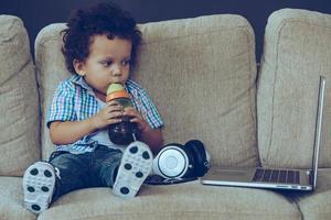 Enjoying favorite catoons. Front view of little African baby boy drinking from baby bottle and looking at his laptop while sitting on the couch at home photo