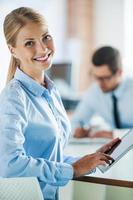 Working hard to become successful. Smiling young businesswoman in formalwear holding digital tablet and looking at camera while her male colleague working in the background photo