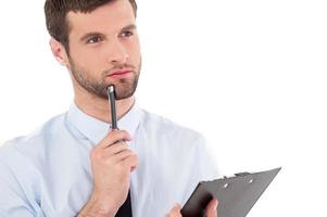 Looking for fresh ideas. Thoughtful young man in formalwear holding clipboard and touching his chin with pen while standing isolated on white background photo