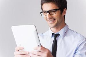 examinando un aparato nuevo. joven feliz con camisa y corbata mirando una tableta digital y sonriendo mientras se enfrenta a un fondo gris