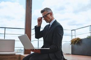 Confident mature man working on laptop while standing on the rooftop terrace photo