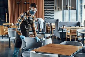 Young men in protective face shields and aprons arranging furniture in restaurant photo