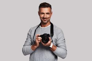 Creative man. Good looking young man holding digital camera and smiling while standing against grey background photo