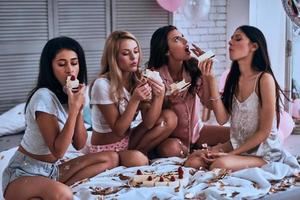 torta deliciosa. cuatro hermosas mujeres jóvenes en pijama comiendo pastel mientras tienen una fiesta de pijamas en el dormitorio foto