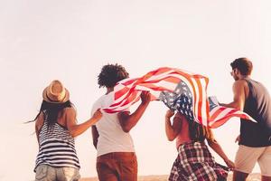 amigos con bandera americana. vista trasera de cuatro jóvenes que llevan la bandera americana mientras corren al aire libre foto