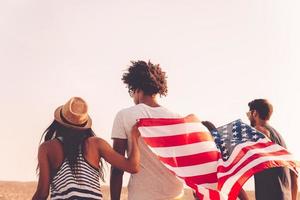 Enjoying freedom. Rear view of four young people carrying american flag while running outdoors photo