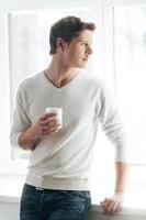 Enjoying good morning. Cheerful young man drinking coffee and looking at camera while standing against window photo