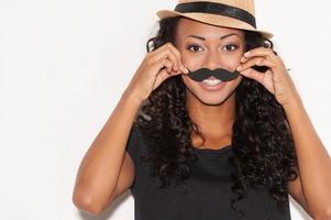 Having fun with face mustache. Cheerful young African woman in funky hat holding fake mustache on her face and looking at camera while standing against white background photo