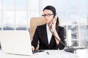 Businesswoman at work. Confident young business woman talking on the phone and using computer while sitting at her working place photo