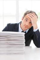 Depressed and overworked. Depressed young man in shirt and tie looking at camera and holding head in hand while sitting at the table with stack of documents laying on it photo