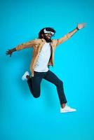 Full length of playful young African man wearing virtual reality glasses while hovering against blue background photo