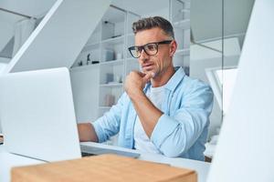 Confident mature man working on laptop while sitting at the office desk photo