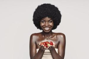 Portrait of beautiful young African woman holding strawberry and smiling while standing against gray background photo