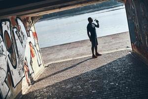 hora de refrescarse. Longitud total de un joven africano con ropa deportiva bebiendo agua mientras hace ejercicio cerca del río al aire libre foto