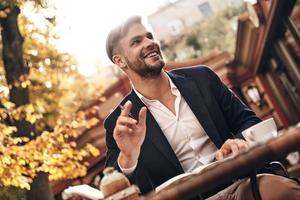 su negocio es su vida. un joven apuesto con ropa informal elegante sonriendo y mirando hacia otro lado mientras se sienta en un restaurante al aire libre foto