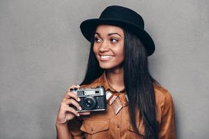 hermosa fotógrafa bella joven africana alegre sosteniendo una cámara de estilo retro y mirando hacia otro lado con una sonrisa mientras se enfrenta a un fondo gris foto