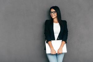 Young business expert. Attractive young woman in smart casual wear and eyeglasses carrying laptop and smiling while standing against grey background photo