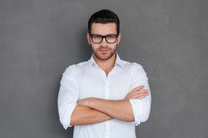 Confident and successful. Handsome young man buttoning his white shirt and looking at while standing against grey background photo