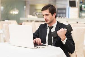 Sports and business. Confident young man in formalwear sitting at laptop and holding a dumbbell photo