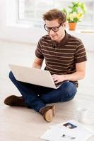 Surfing web at home. Top view of handsome young man in glasses working on laptop while sitting on the floor at his apartment photo