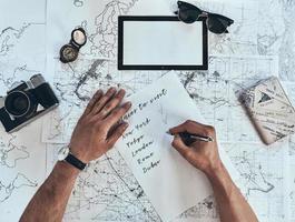 Making a list. Close up top view of man writing down checklist with sunglasses, photo camera, compass, passport lying on map around