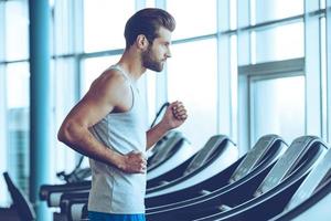 Running fast. Side view of young man in sportswear running on treadmill at gym photo