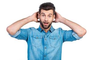 What a surprise Surprised young handsome man in jeans shirt holding hands behind head and keeping mouth open while standing against white background photo