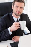 Taking a coffee break. Top view of confident young man in formalwear drinking coffee and smiling while sitting at his working place photo