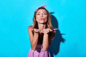 Sending all her love. Beautiful young woman blowing a kiss and looking at camera while standing against blue background photo