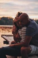 Too good to be true. Happy young couple embracing and smiling while sitting on the pier near the lake photo