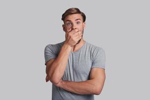 Feeling surprised. Shocked young man making a face and covering mouth with hand while standing against grey background photo