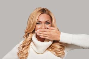 Stop gossiping Playful young smiling woman looking at camera and covering mouth with hand while standing against grey background photo