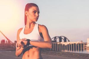 Ready to run now. Beautiful young woman in sports clothing wearing smartwatch and looking away while standing on the bridge photo